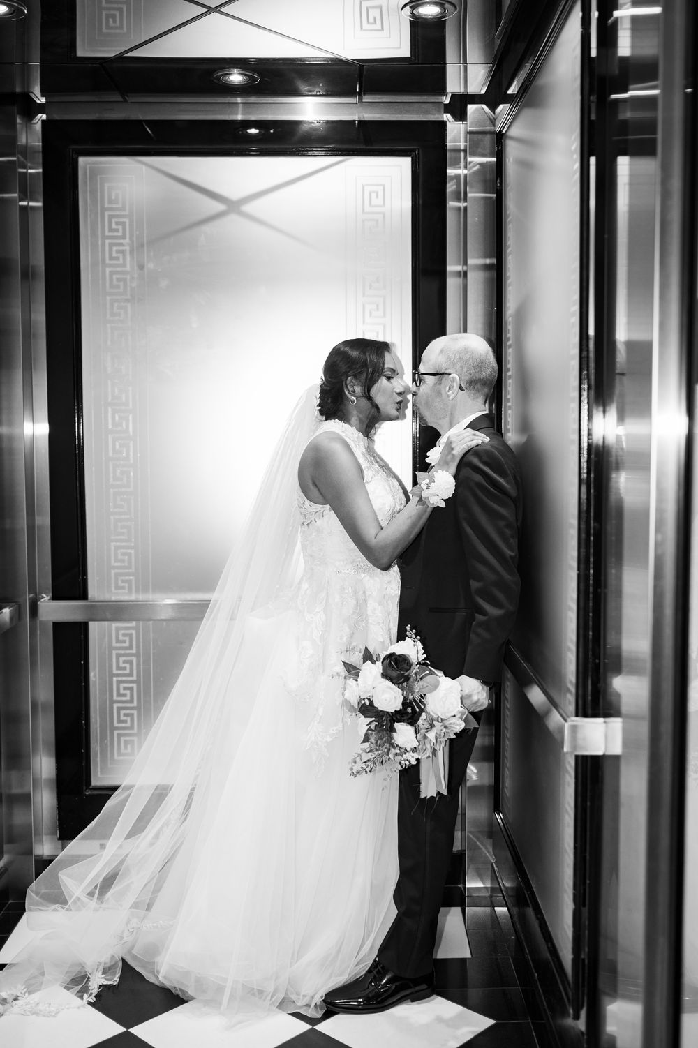 Black and white wedding photo of bride and groom sharing a kiss in an elegant elevator, bride holding a bouquet.