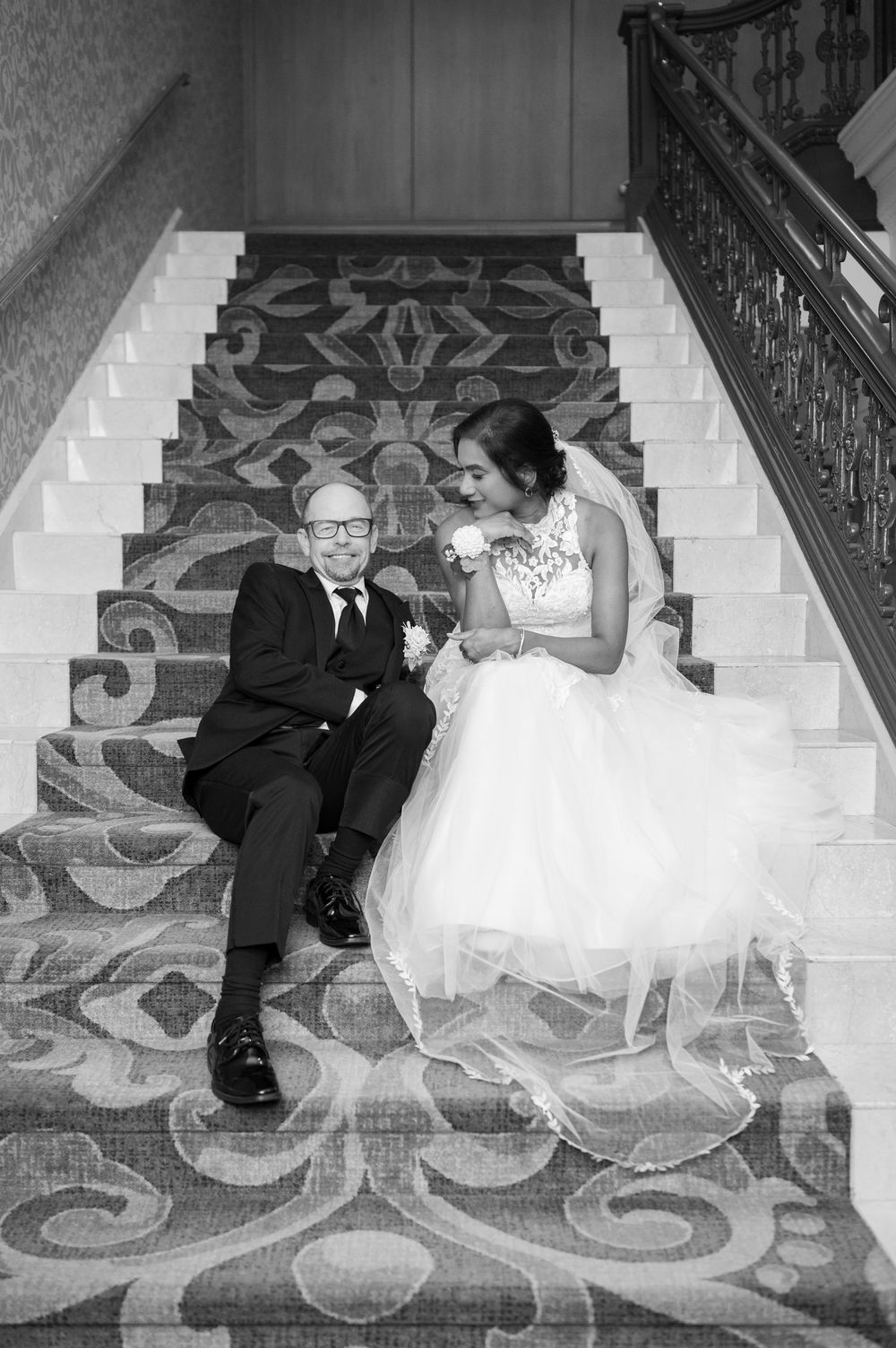 Black and white photo of a bride and groom sitting together on ornate carpeted stairs, smiling at each other.