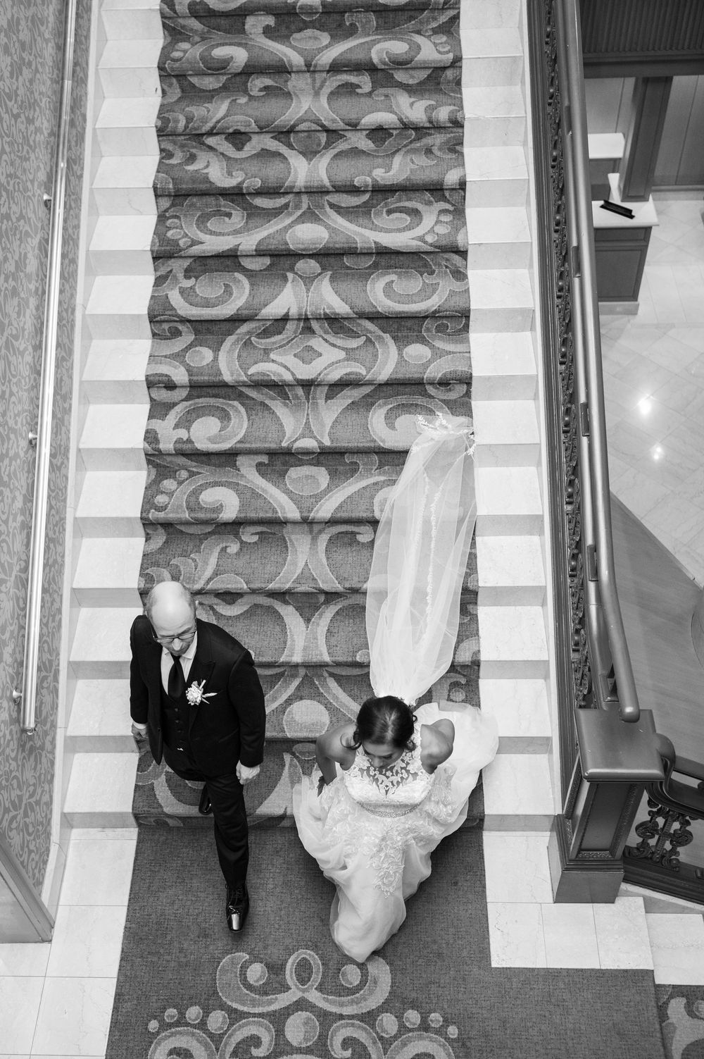 Black and white aerial view of bride and groom descending ornate patterned staircase, wedding gown train trailing behind.