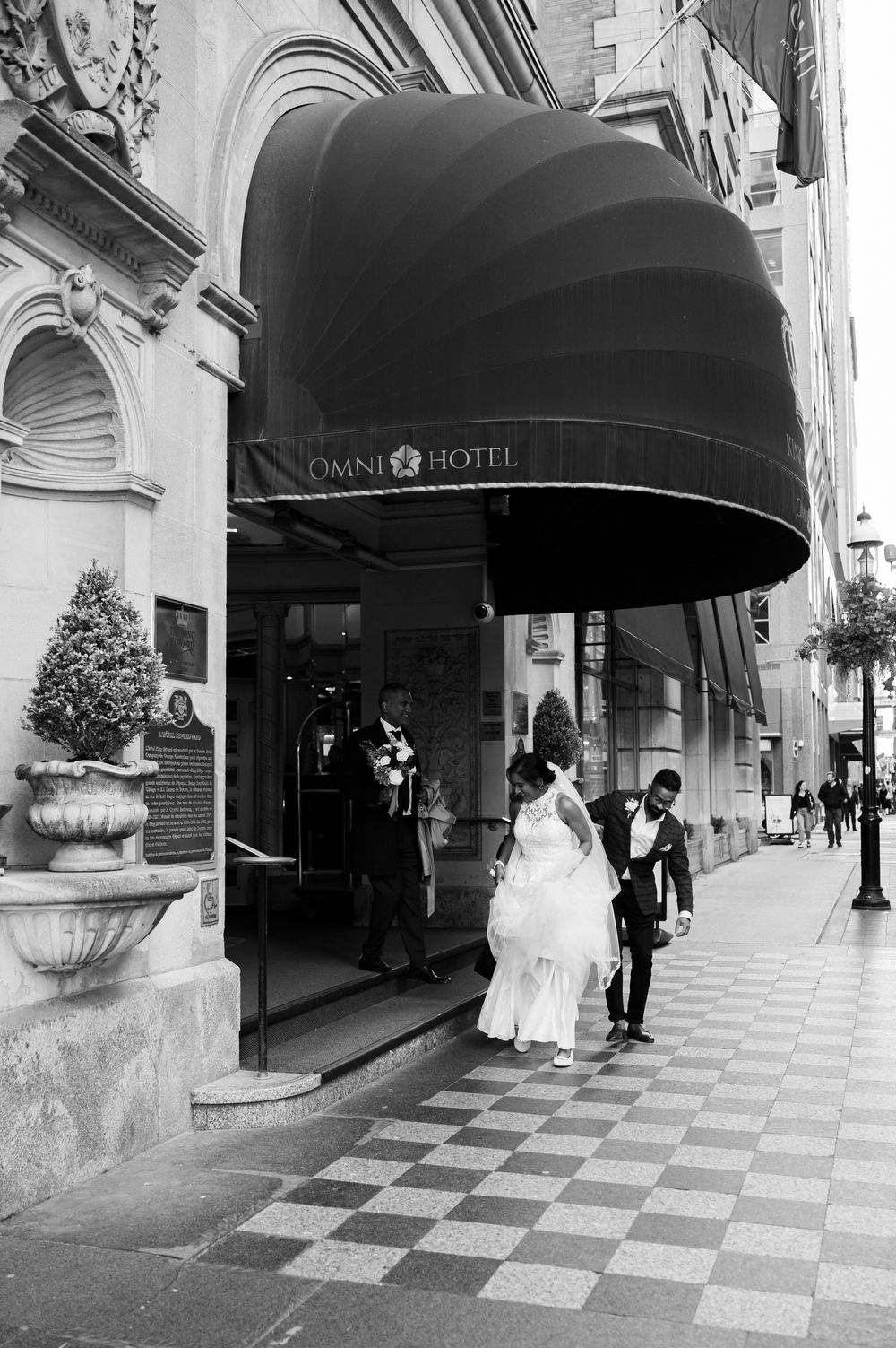 Bride and groom exiting Omni Hotel entrance on checkered sidewalk in elegant wedding attire.