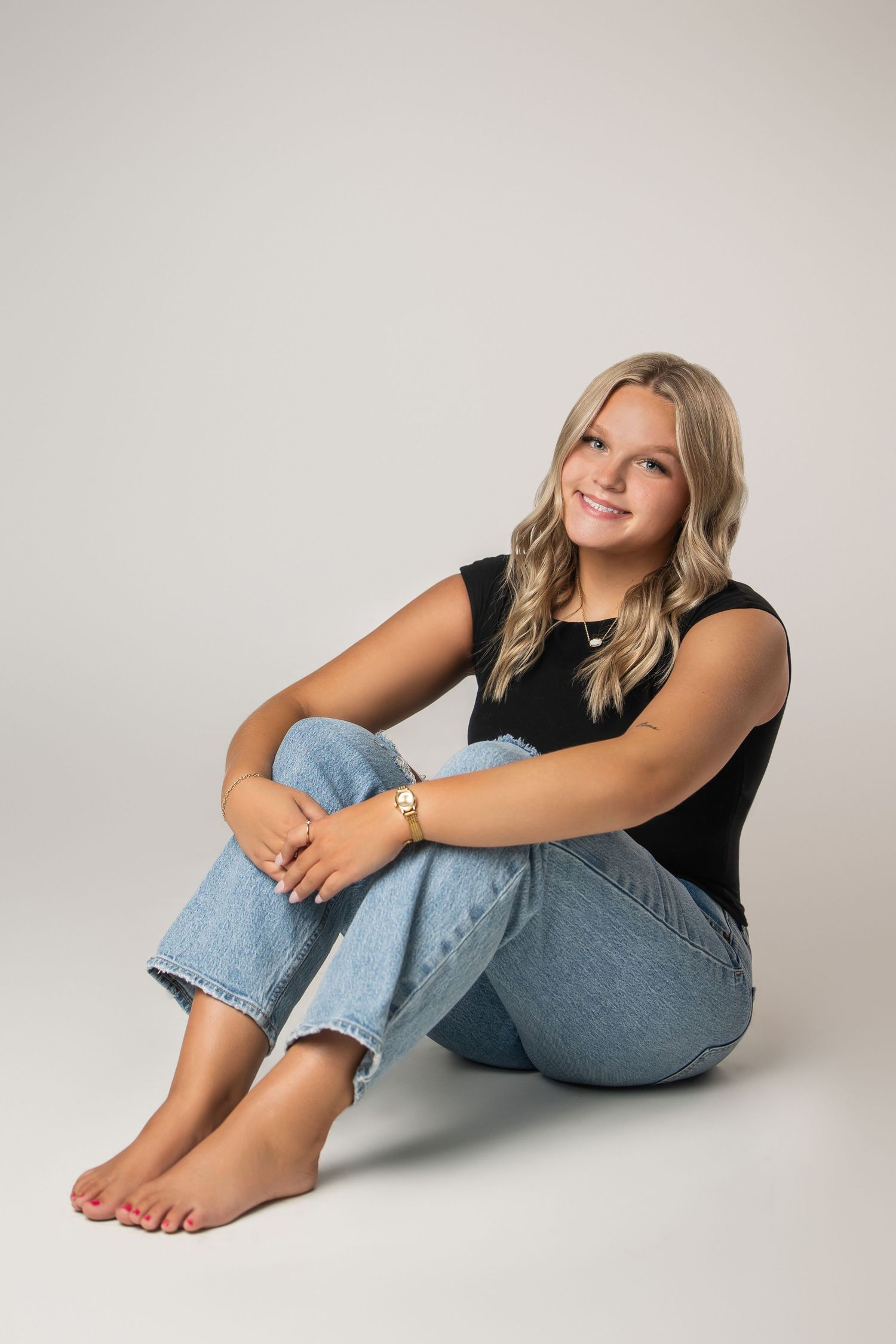Smiling blonde teenage girl sitting barefoot on studio floor wearing black top and light blue jeans with gold bracelet.