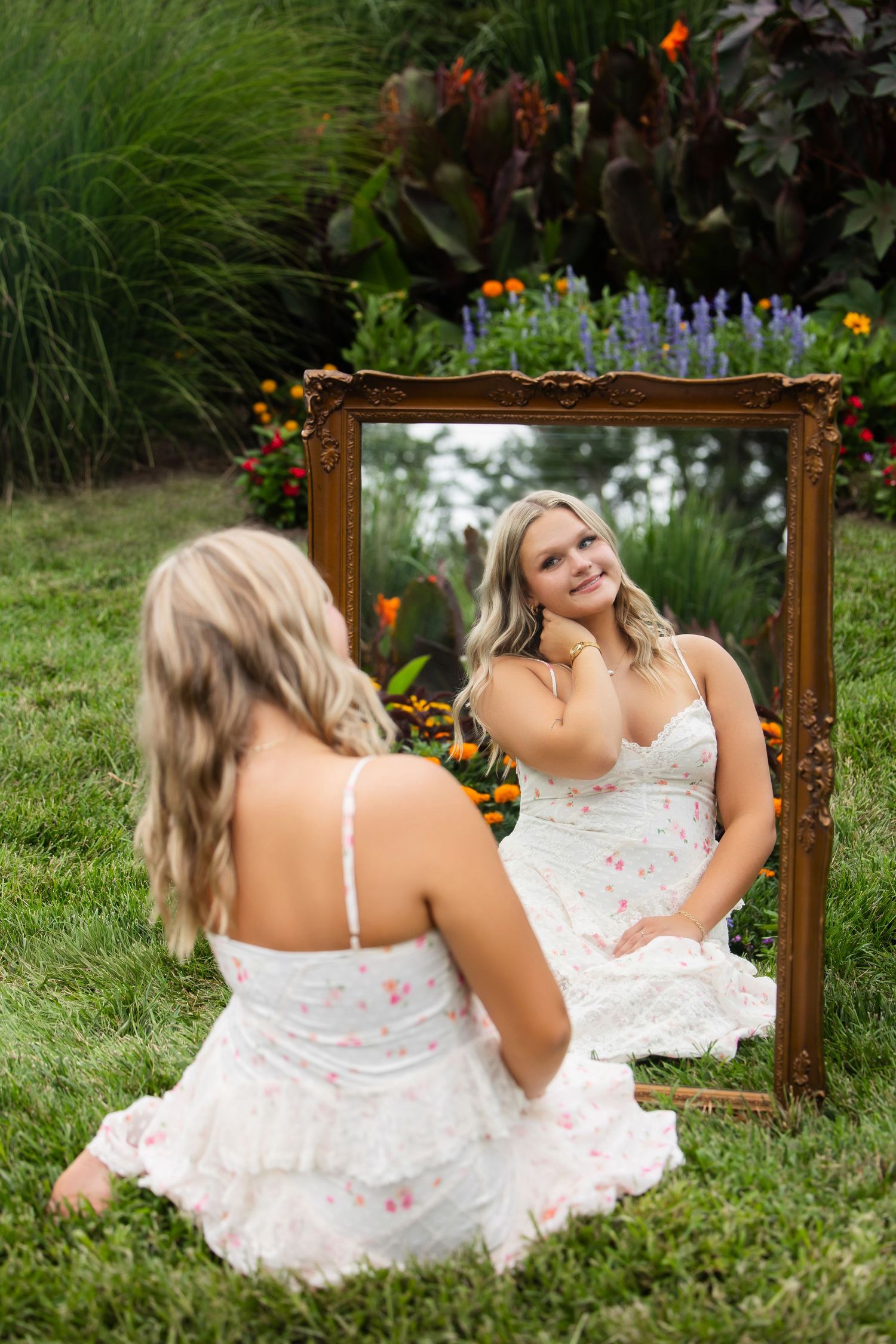 Young blonde woman in floral dress sitting on grass, smiling at her reflection in an ornate gold mirror outdoors.