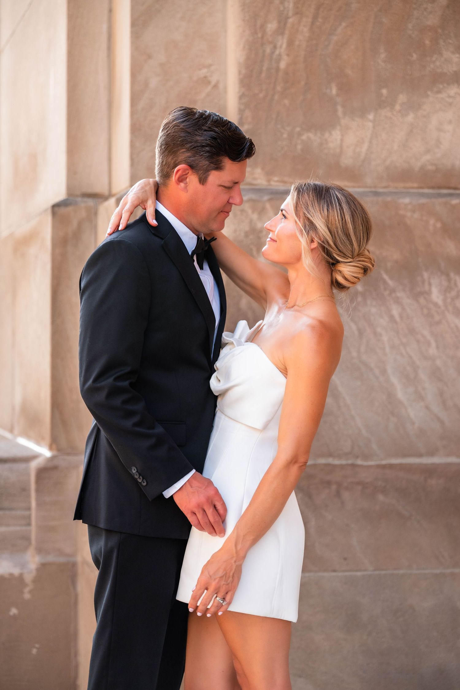 Elegant couple in black tuxedo and white strapless mini dress embracing near stone columns.