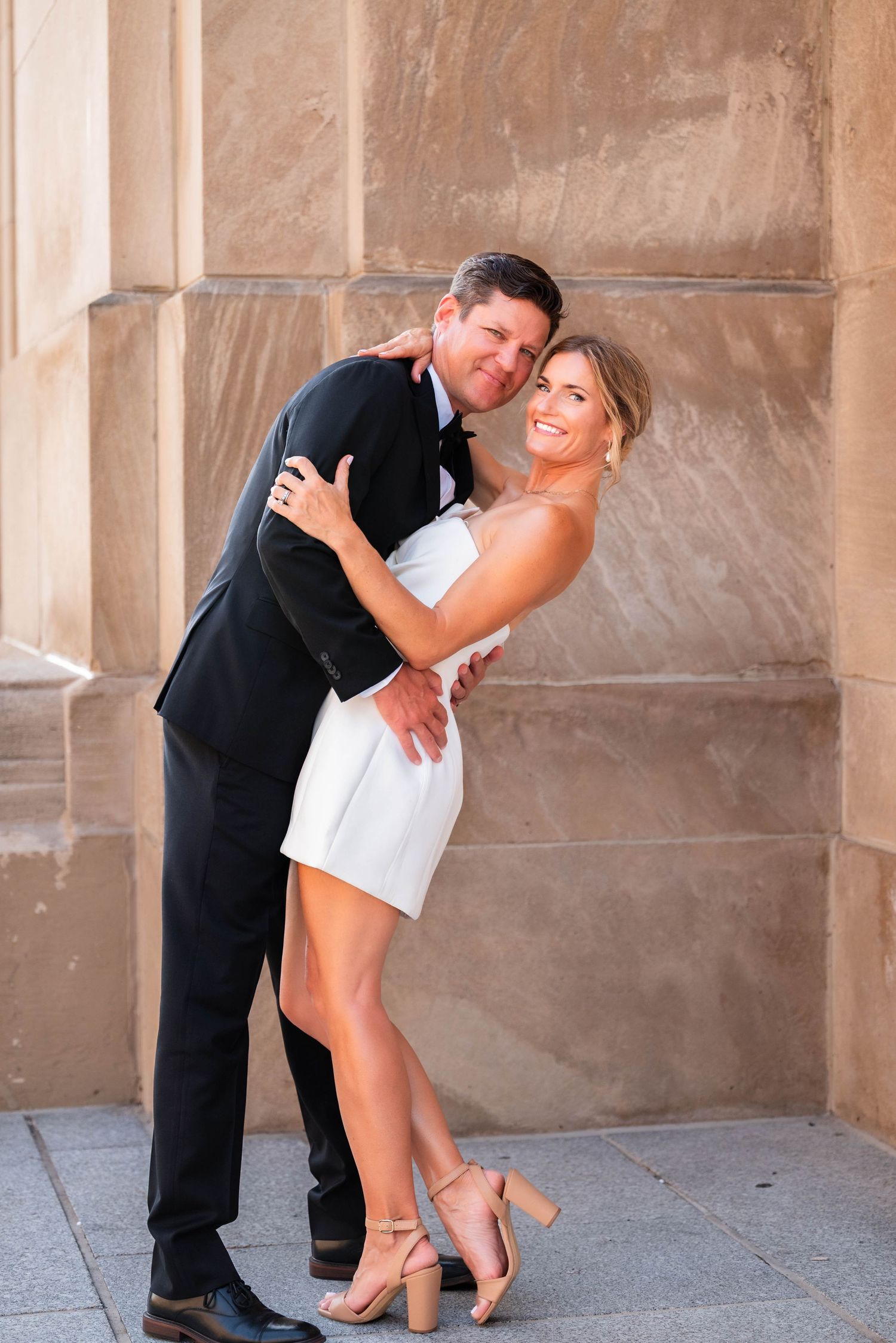Happy couple in wedding attire pose romantically against a stone wall, bride wearing white mini dress.