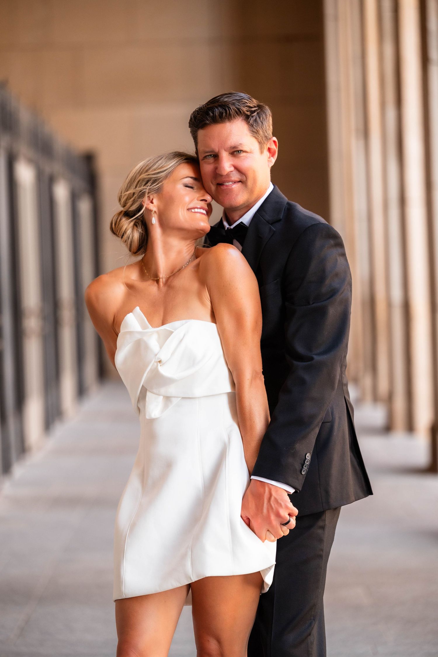 Bride in white strapless mini dress and groom in black tuxedo embrace among elegant stone columns.