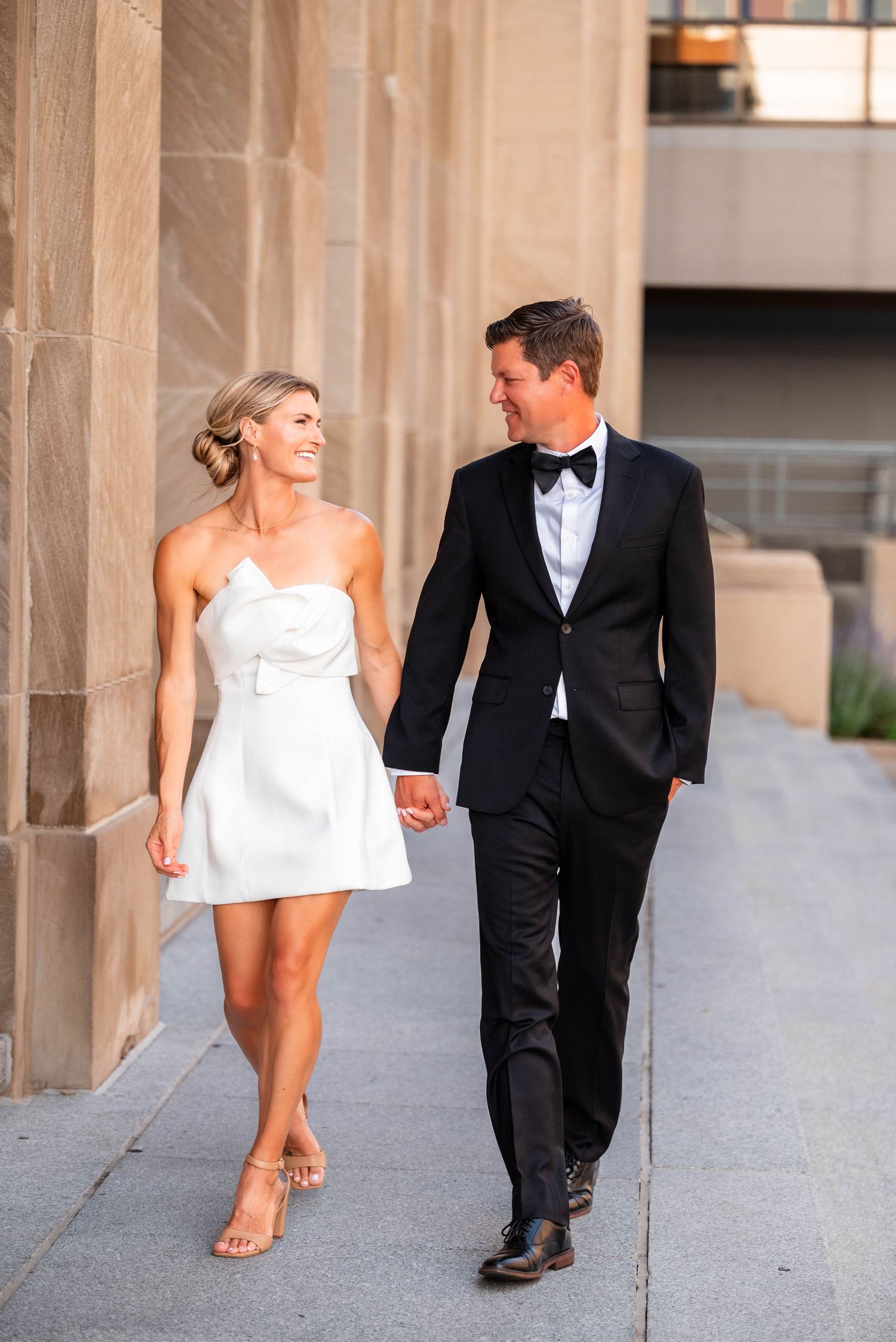 Couple in wedding attire walking hand in hand, woman in white mini dress, man in black tuxedo, smiling at each other.