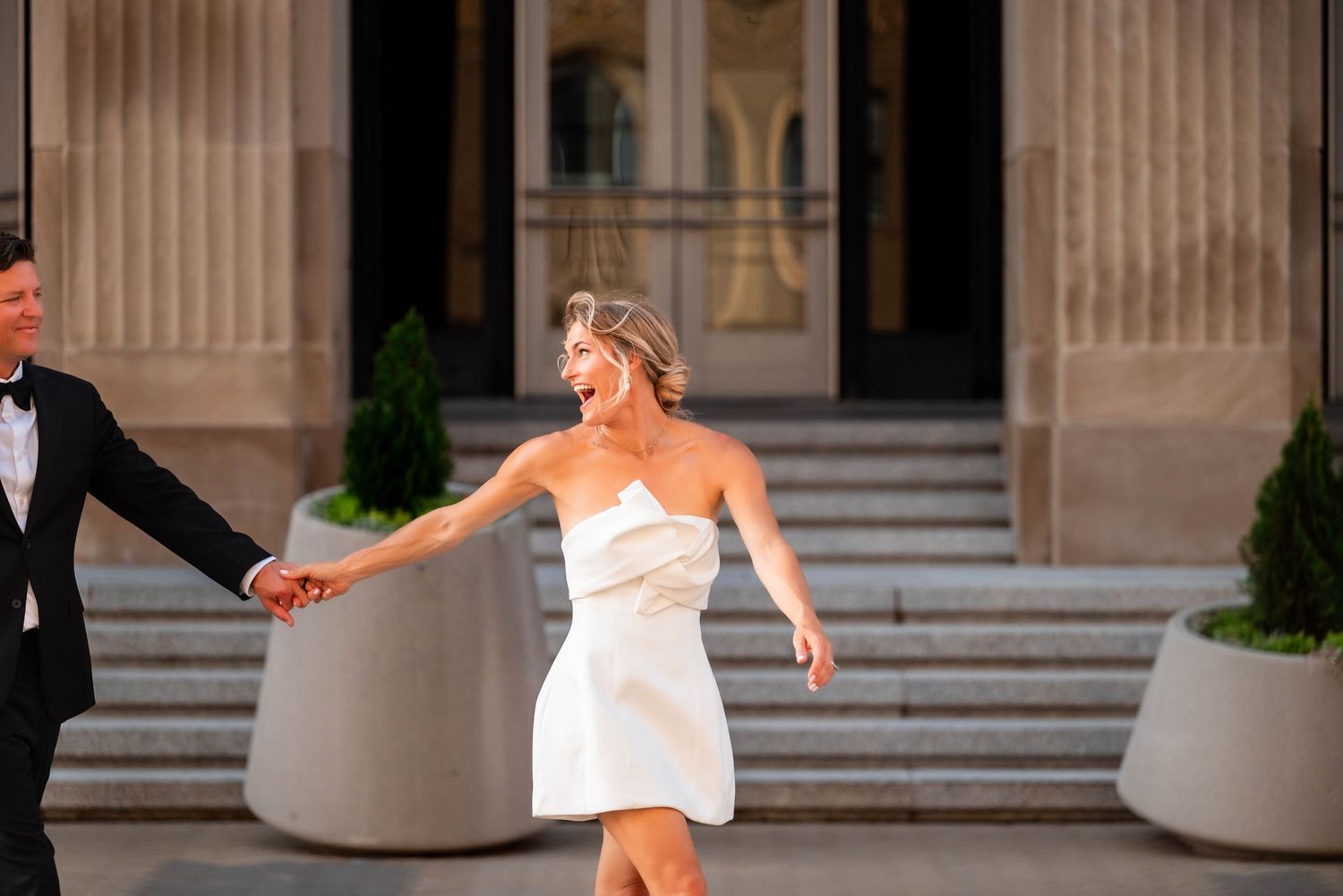 Joyful bride in white mini dress holding hands with groom in tuxedo outside elegant building.