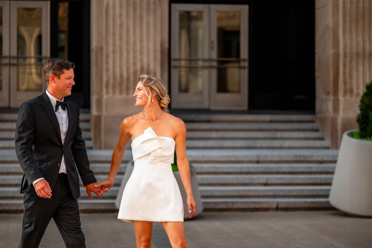 Couple in formal wedding attire holding hands outside stone building, woman in white mini dress, man in black tuxedo.