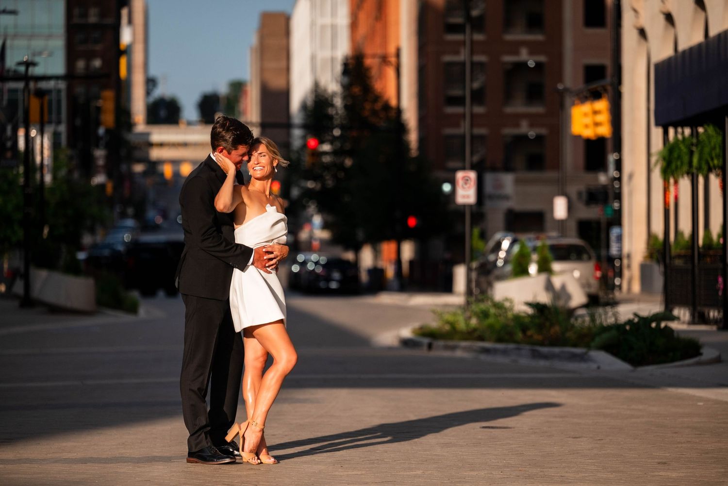 Couple sharing a romantic kiss on a sunny urban street corner, woman in white dress, man in dark suit.