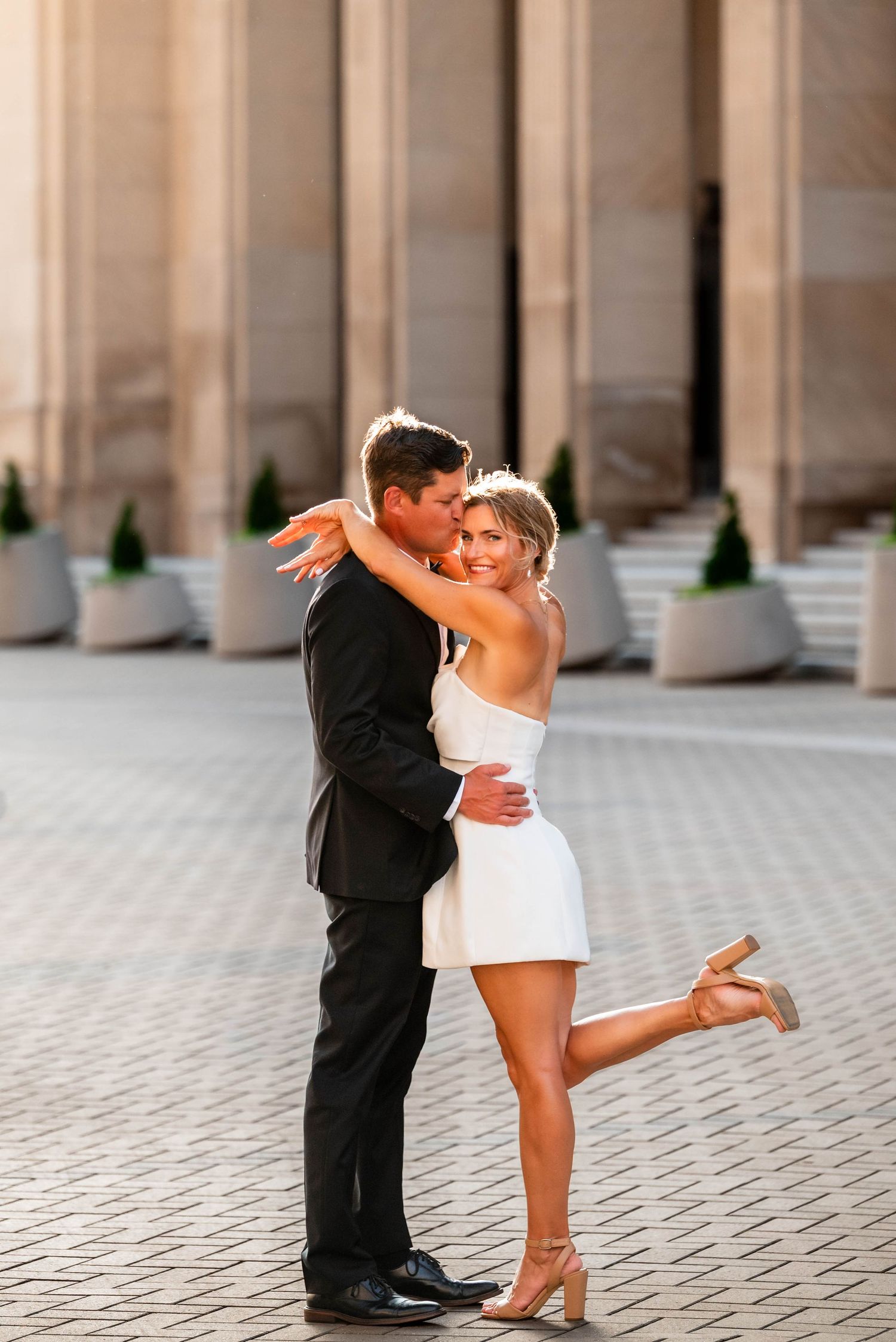 Couple sharing a romantic kiss outdoors, bride in white mini dress and groom in black suit, golden hour lighting.