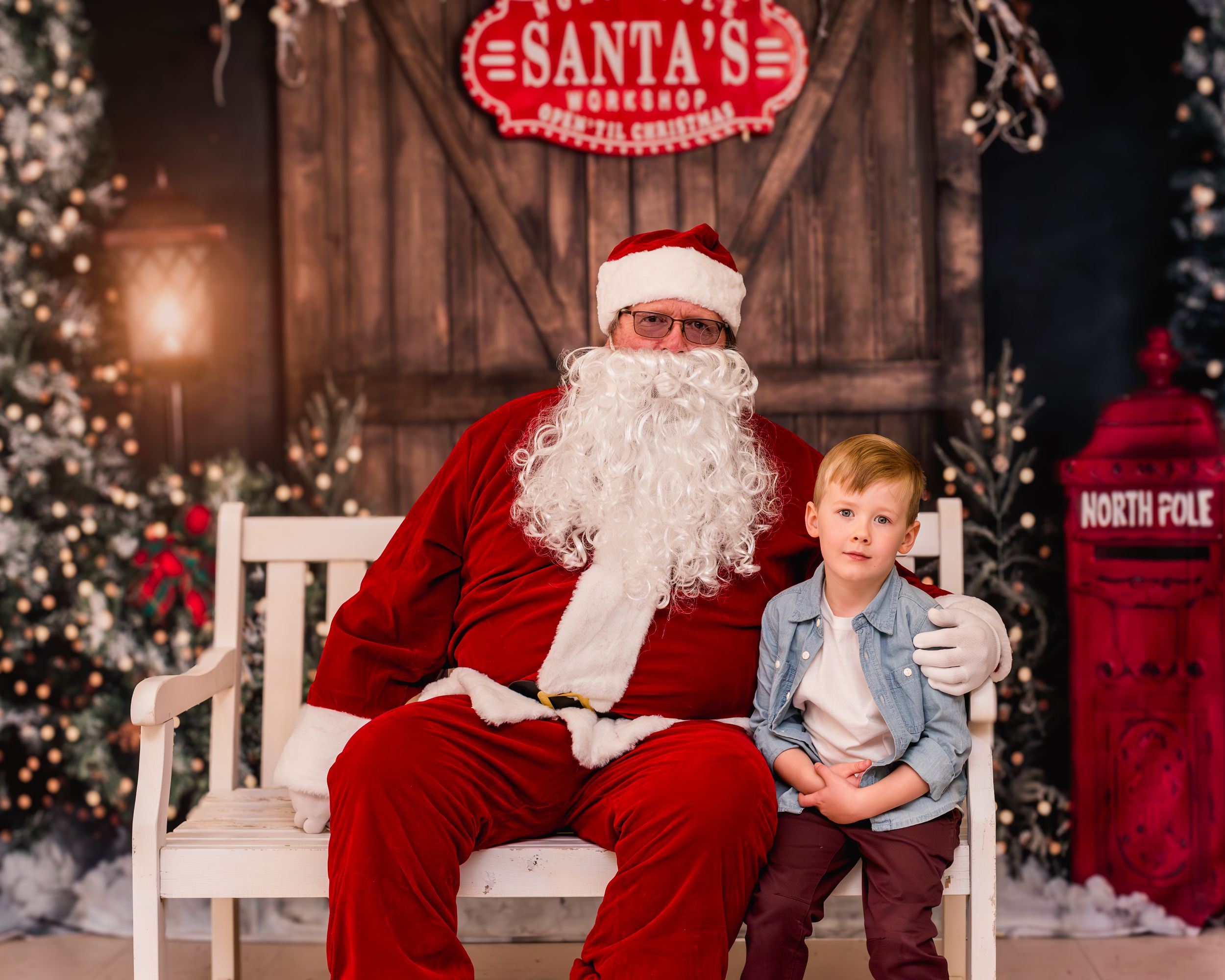 Two children sit with Santa Claus in his workshop, surrounded by festive Christmas decorations and trees.