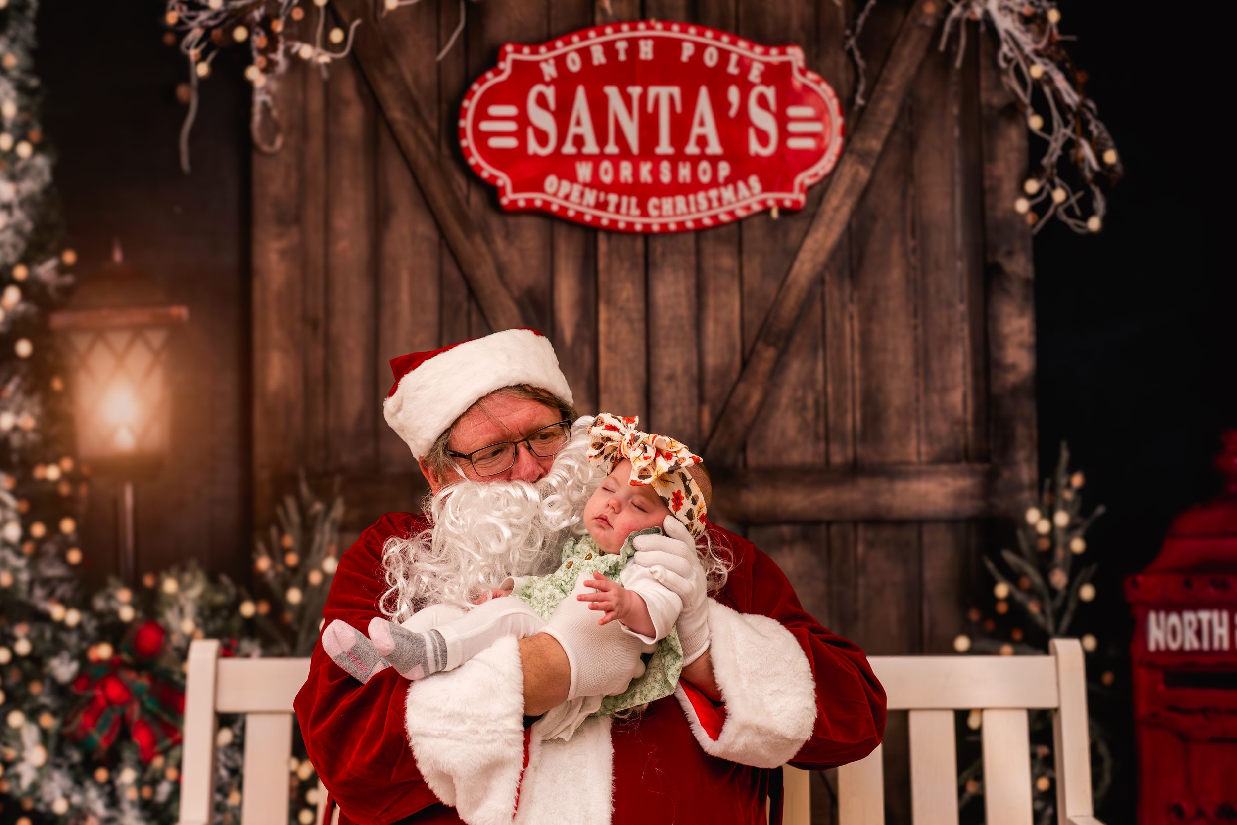 Two children sit with Santa Claus in his workshop, surrounded by festive Christmas decorations and trees.