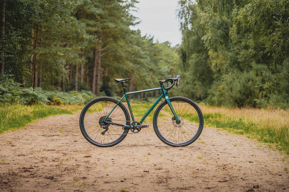 A teal gravel bike parked on a sandy forest trail surrounded by lush green trees and vegetation.