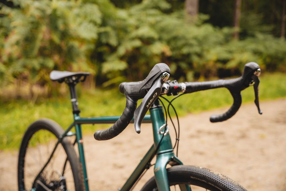 Close-up of a green gravel bike handlebar with drop bars and brake levers, set against a lush forest background.