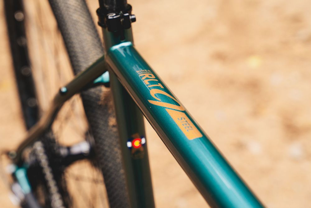 Close-up of a teal green bicycle frame with gold logo detailing, showcasing sleek tube design against a sandy background.