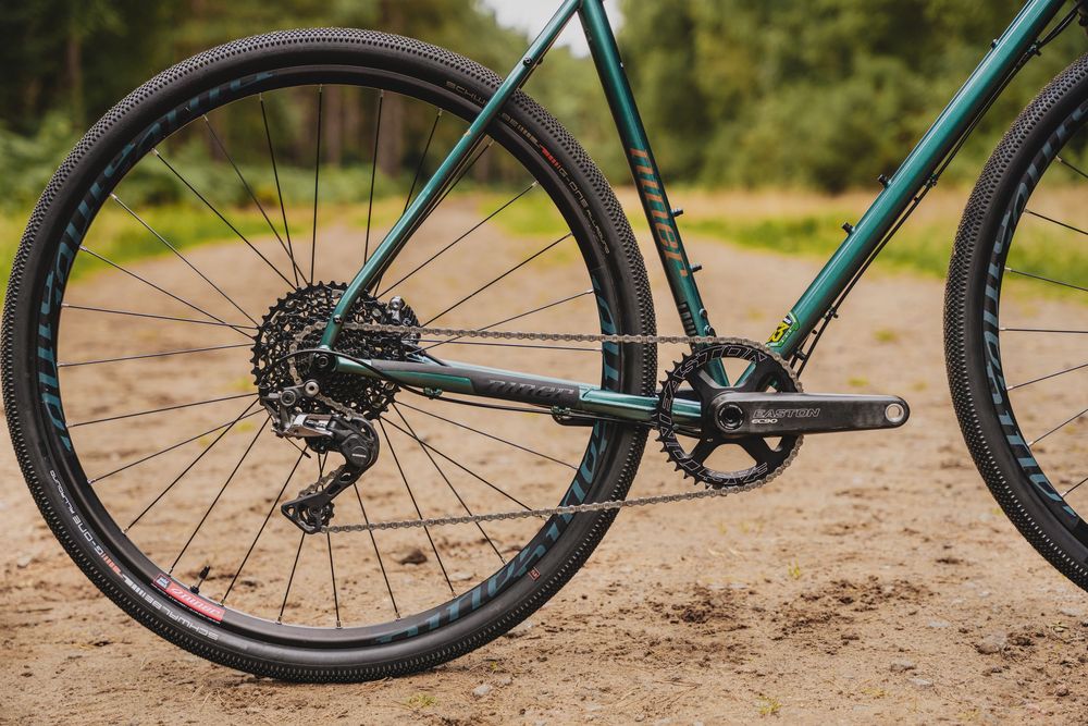 Close-up of a green gravel bike rear wheel, cassette, derailleur, and chainring on a sandy dirt path.