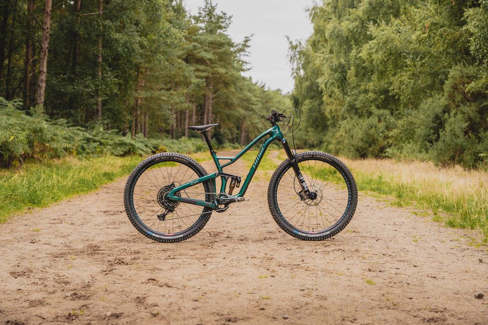 Green full-suspension mountain bike parked on a sandy forest trail surrounded by lush green trees.