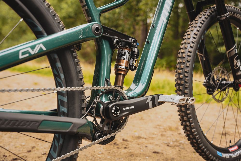 Close-up of a green full-suspension mountain bike frame showing CVA linkage, rear shock, chain, and knobby tire on dirt trail.