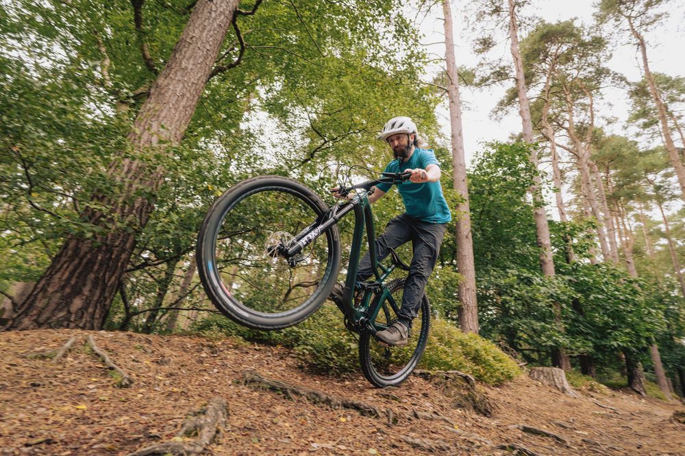 A mountain biker performs a wheelie on a forest trail, wearing a helmet and teal shirt among tall trees.