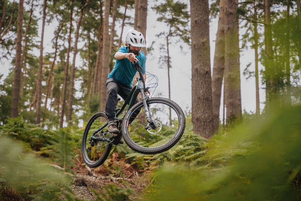 A cyclist performs a wheelie on a mountain bike trail through a lush green forest wearing a white helmet.