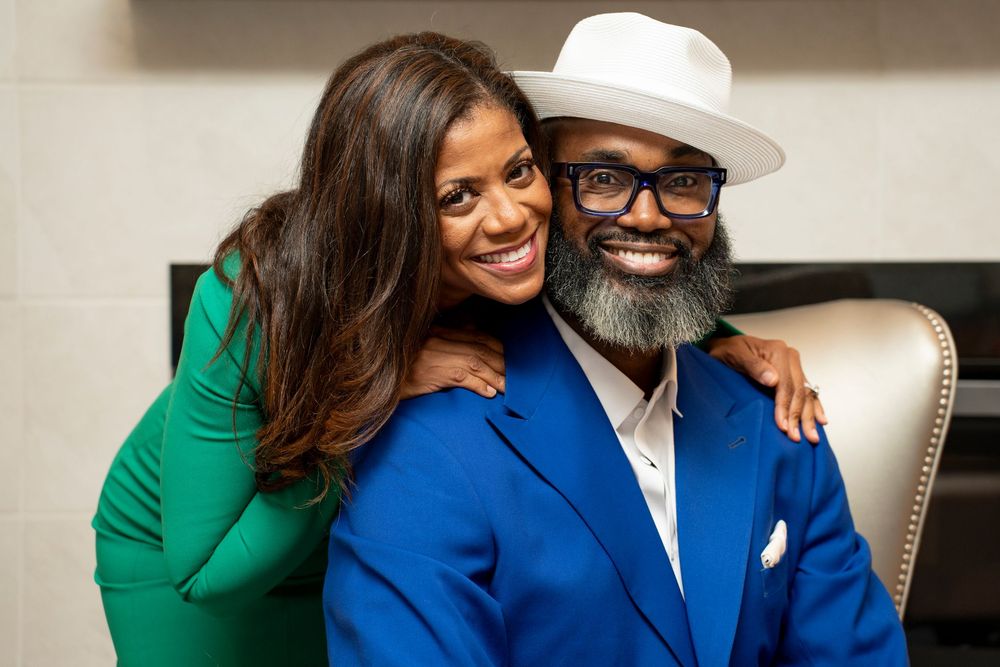 Smiling Black couple posing together, woman in green dress and man in blue suit with white hat.