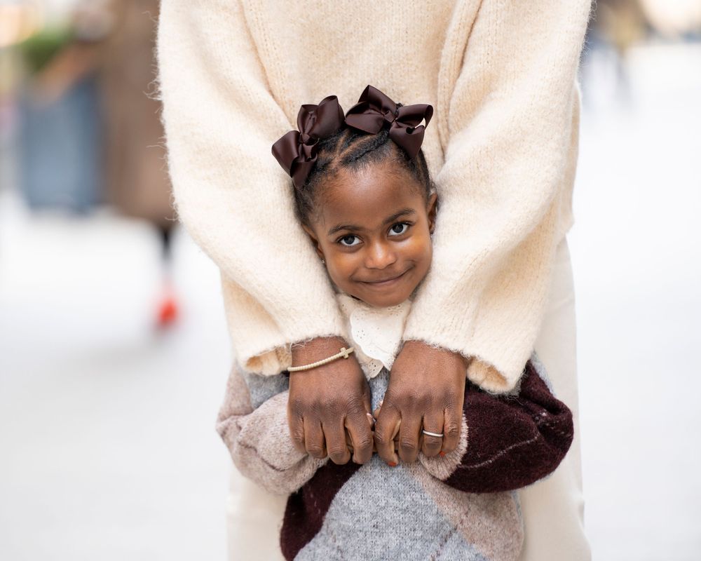Adorable young Black girl with ribbon bows hugging an adult wearing a cream coat.