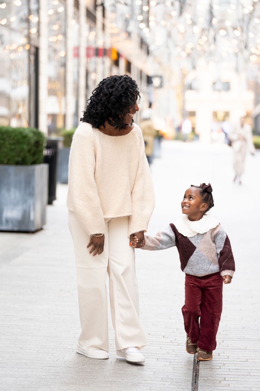Black mother and daughter walking hand-in-hand on a festive city street with holiday lights.