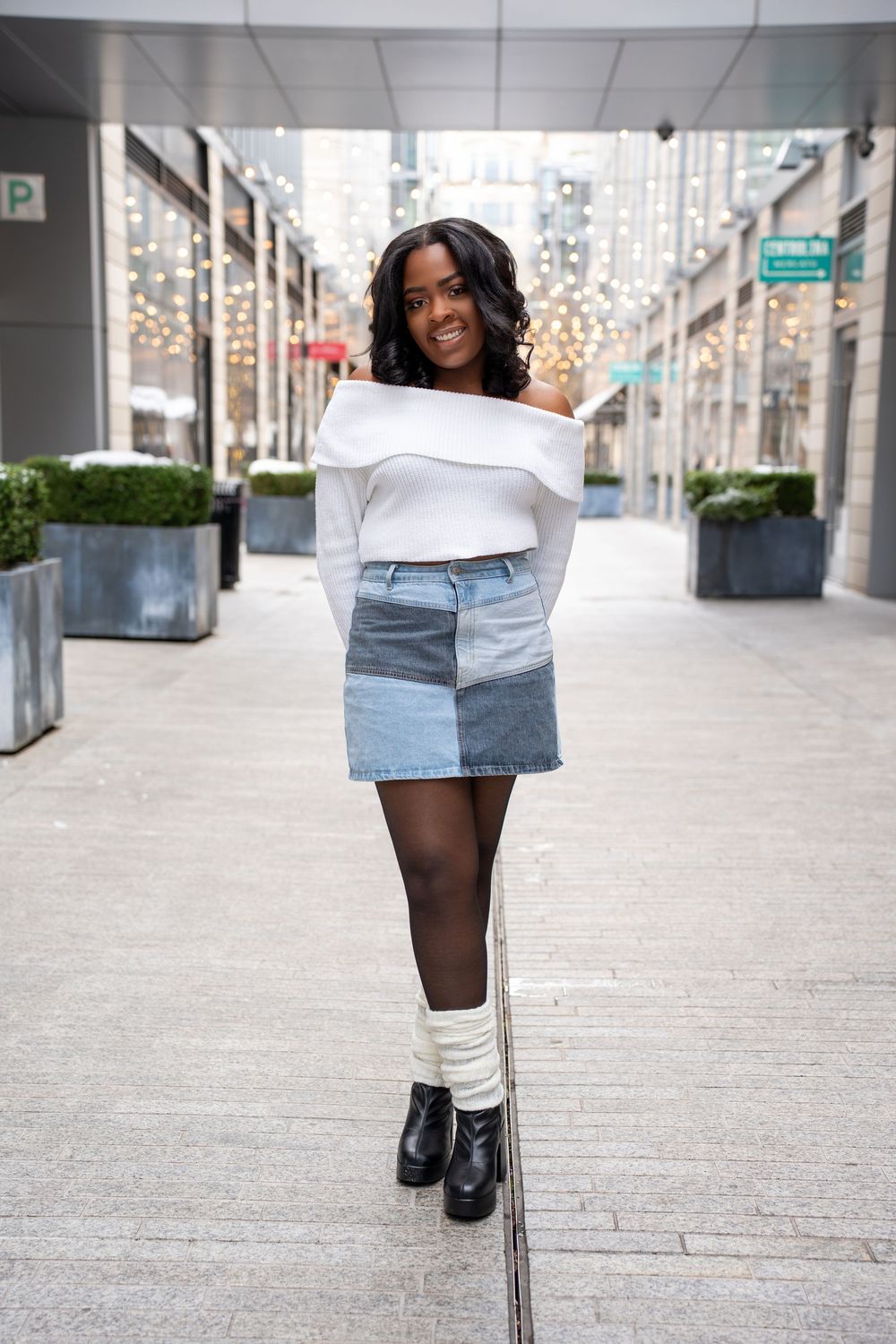 Young Black woman in white off-shoulder top, denim patchwork skirt, and boots on city street.