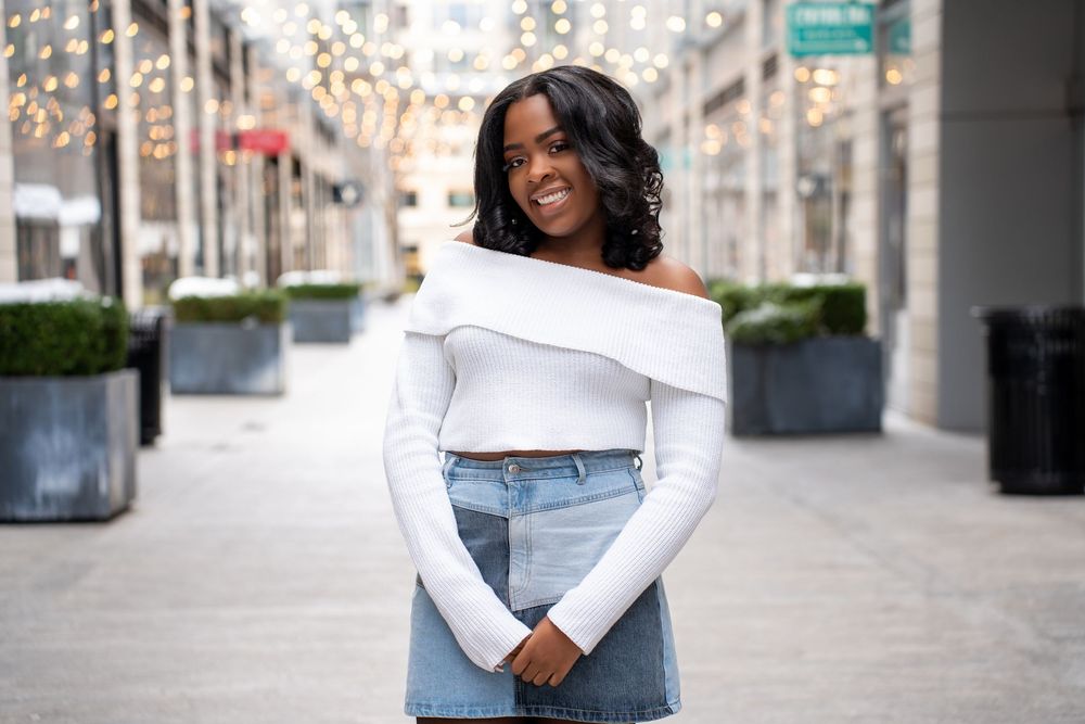 Cheerful Black woman in white off-shoulder crop top and denim skirt smiling on urban walkway.