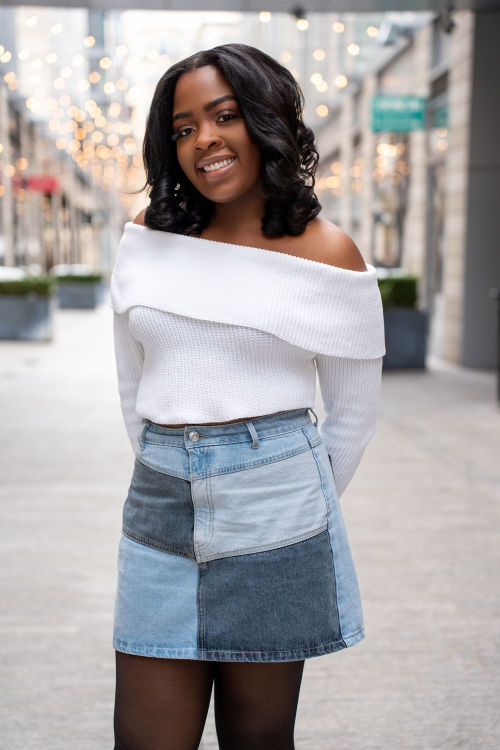 Smiling Black woman wearing white off-shoulder sweater and patchwork denim mini skirt outdoors.