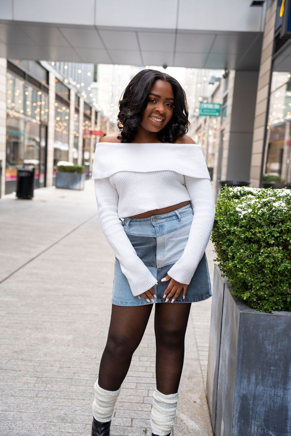 Young Black woman smiling in white off-shoulder crop top, denim patchwork mini skirt, black tights, and leg warmers on city sidewalk.