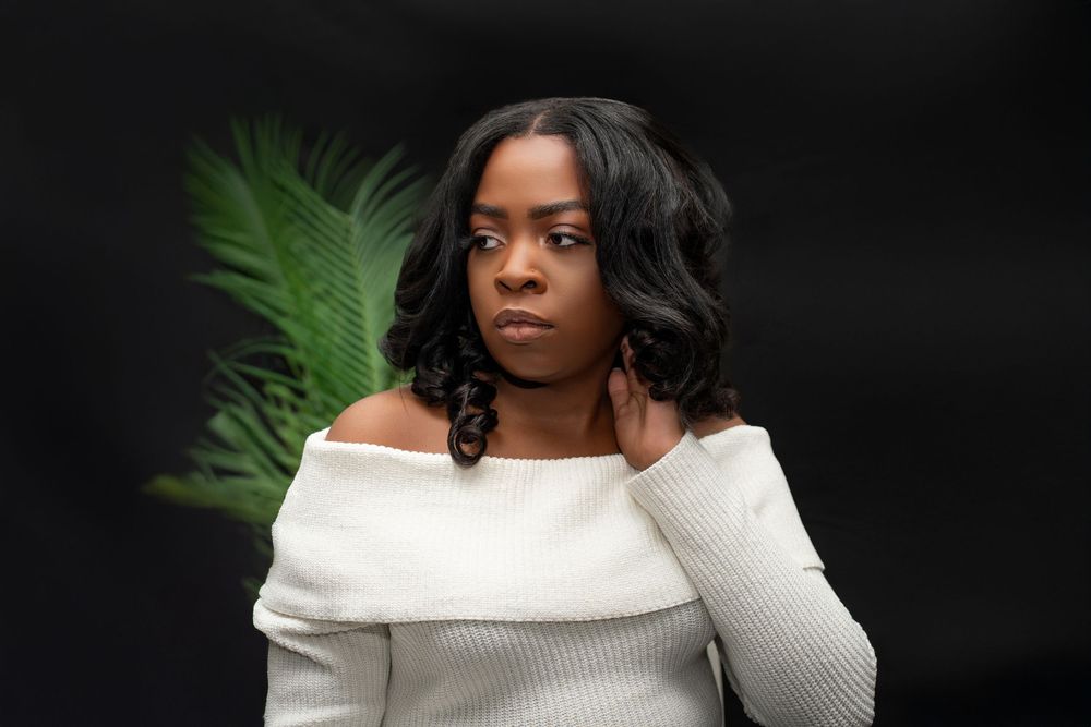 Young Black woman in white off-shoulder ribbed sweater posing against dark background with tropical plant.