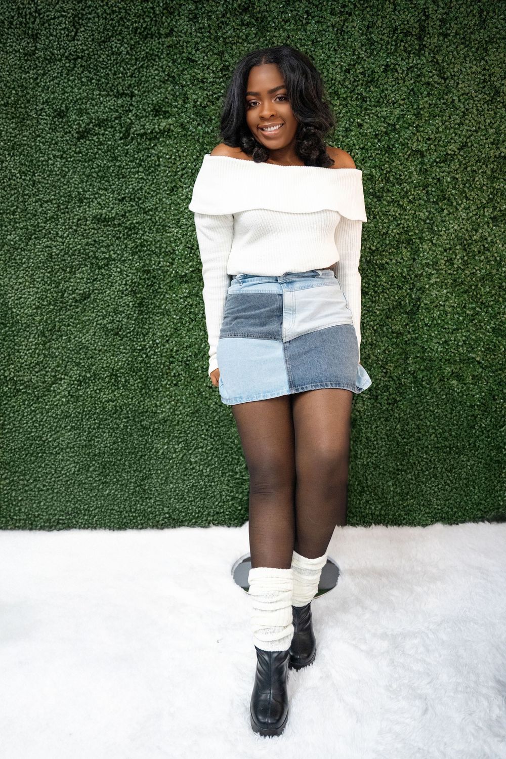 Smiling Black woman in white off-shoulder top and patchwork denim skirt posing against green hedge wall.