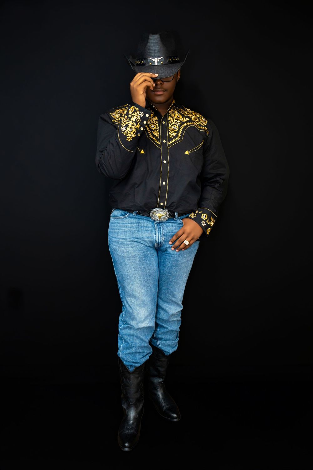 Black man in embroidered western shirt, cowboy hat, jeans, and boots posing on dark background.