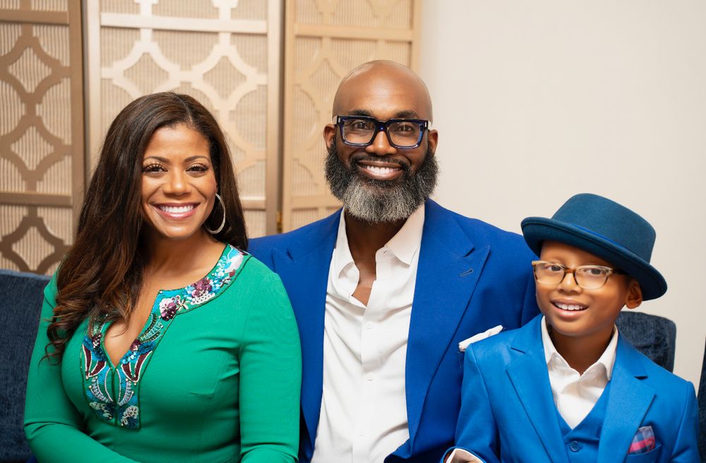 Smiling Black family of three dressed in blue and green outfits posing together indoors.