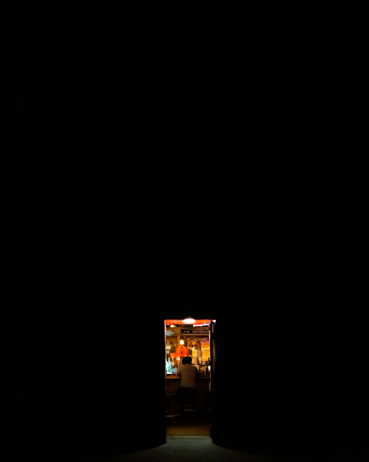A dimly lit doorway glows with warm light revealing a small shop interior against a dark surrounding.