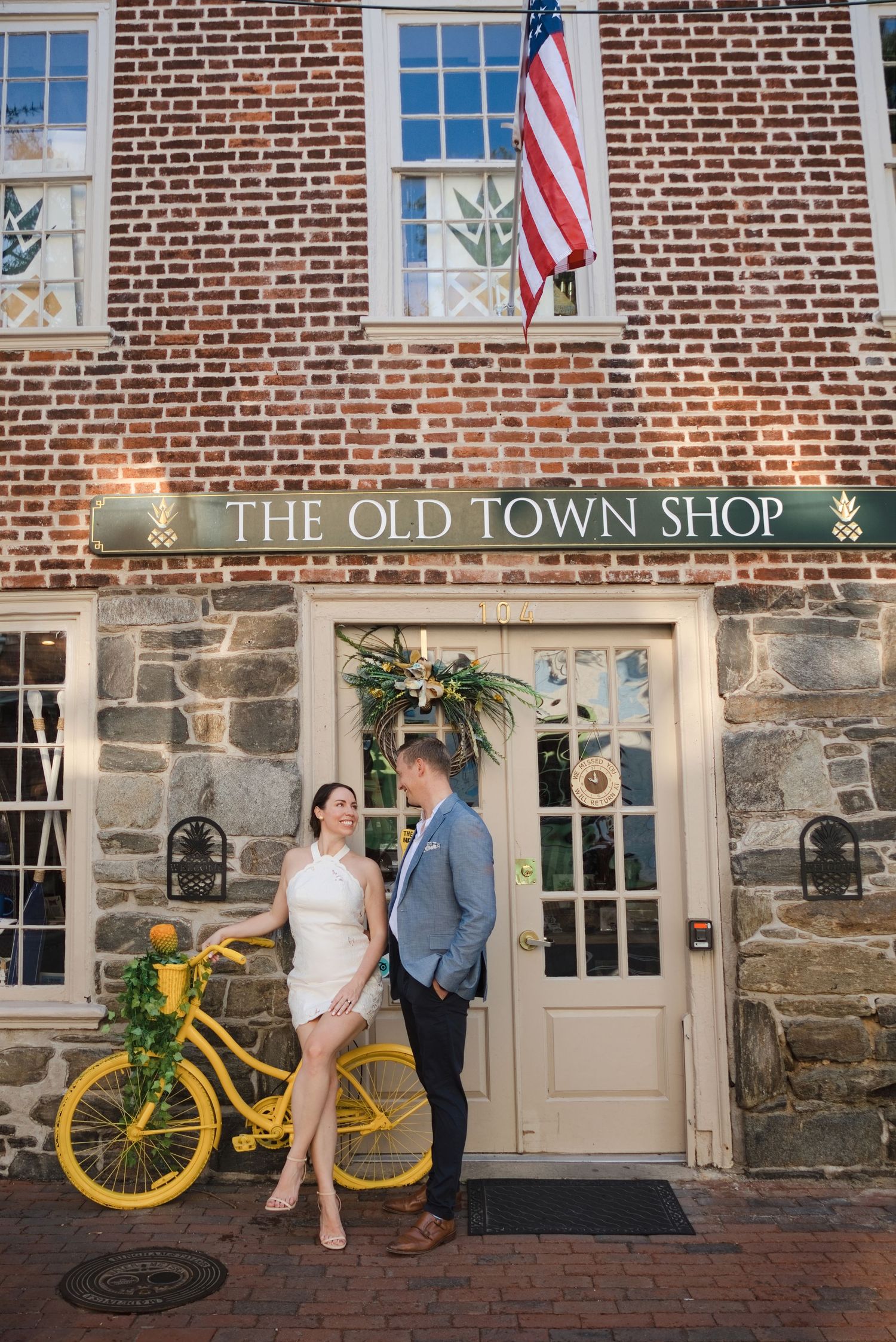 Couple posing in front of The Old Town Shop brick building with a yellow bicycle and American flag.