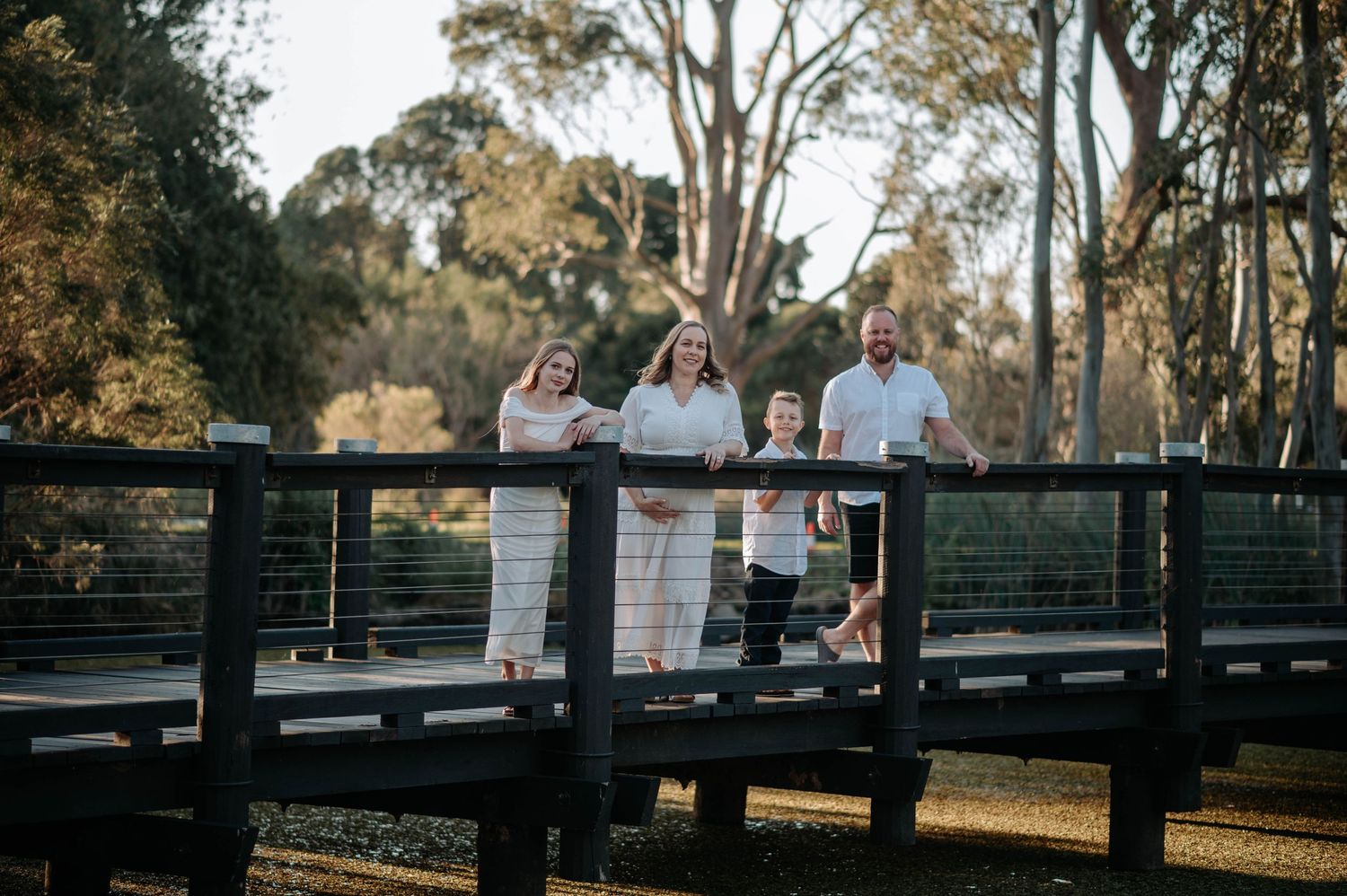 Family of four posing on a wooden bridge surrounded by tall trees in a sunny outdoor park setting.
