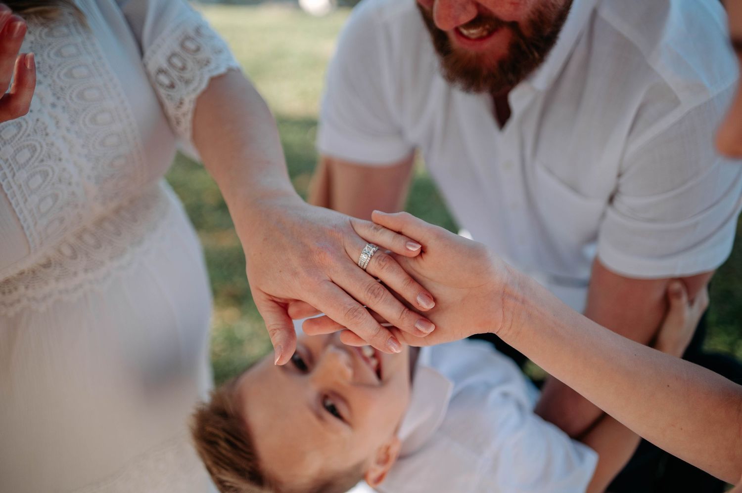 Bride and groom stacking hands with child, showing off wedding ring during outdoor family celebration.