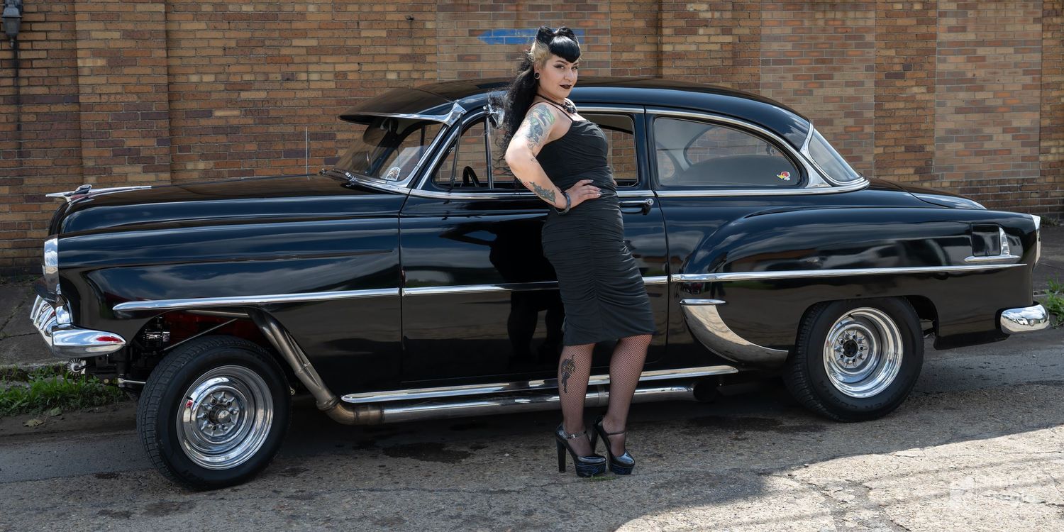 Woman in black dress posing beside a classic black vintage car outdoors near a brick building.