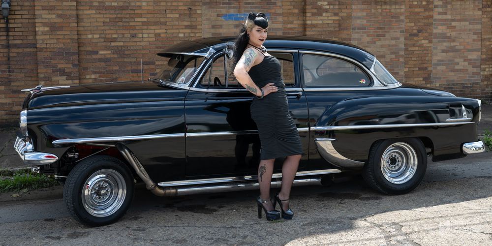 Woman in black dress posing beside a classic black vintage car outdoors near a brick building.