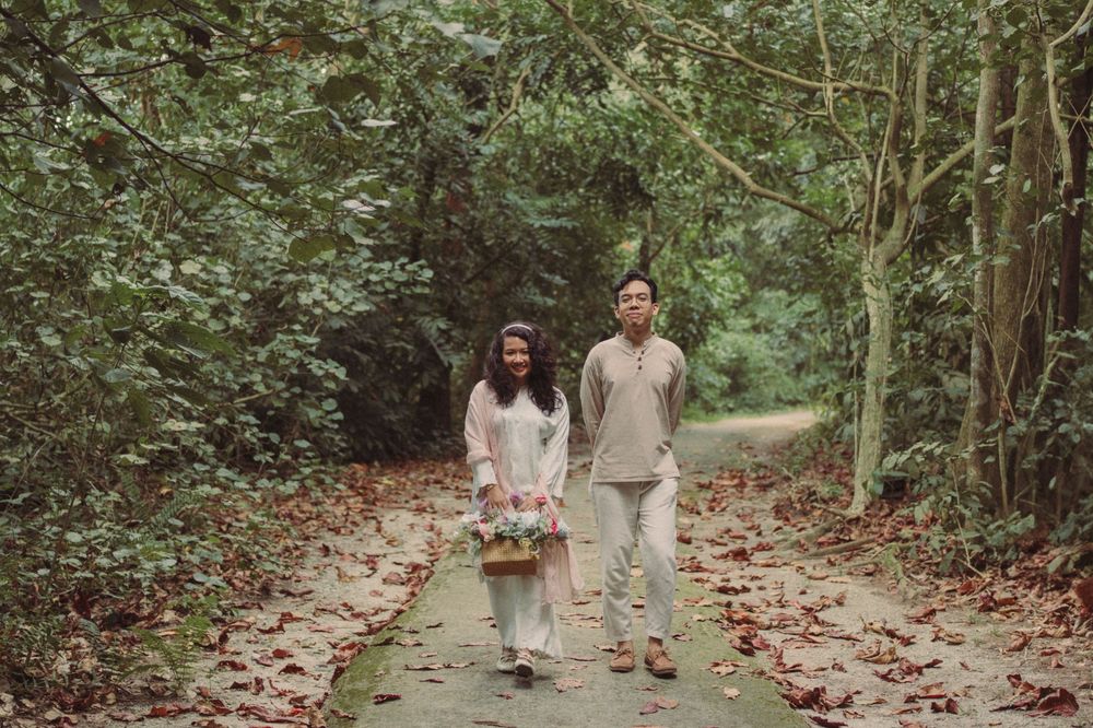 Couple walking hand in hand on a forest path surrounded by lush green trees, carrying a flower basket.