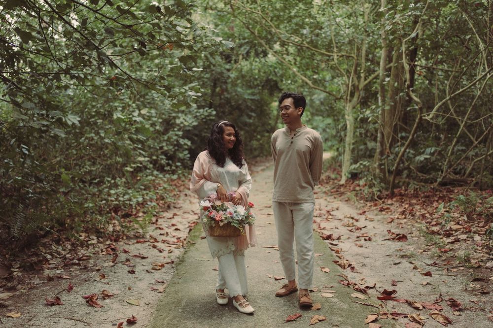 Couple walking on a forest path, woman holding a bouquet, surrounded by lush green trees and fallen leaves.