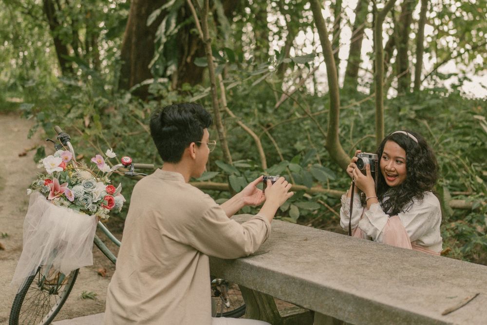 Couple at outdoor picnic table, man photographing smiling woman holding camera, bicycles with flowers in forest background.