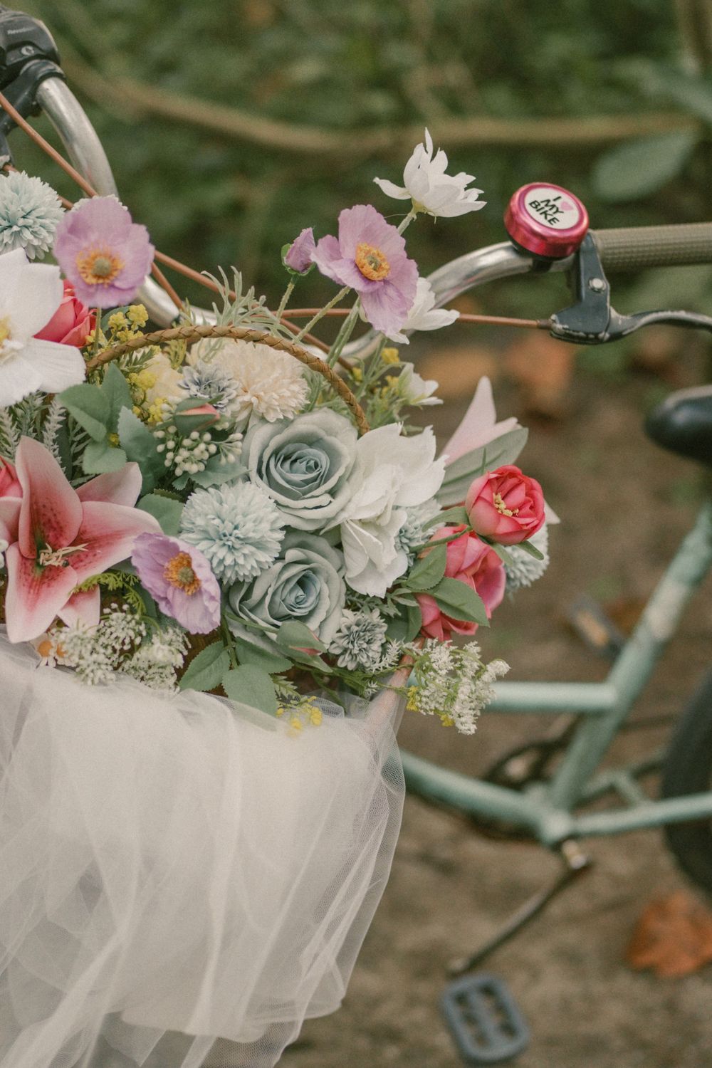 A romantic floral bouquet with pink lilies, roses, and white blooms in a bicycle basket draped with white tulle.