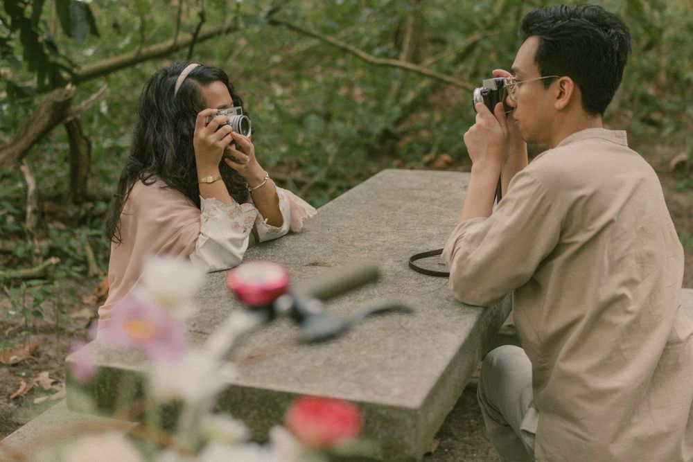 A couple sits at a stone table outdoors, photographing each other with vintage cameras in a lush garden setting.