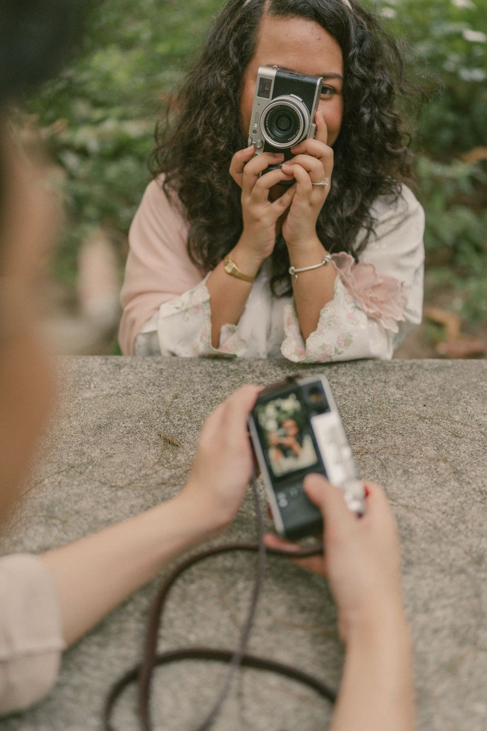 Two women photographing each other with vintage cameras outdoors, surrounded by green foliage.