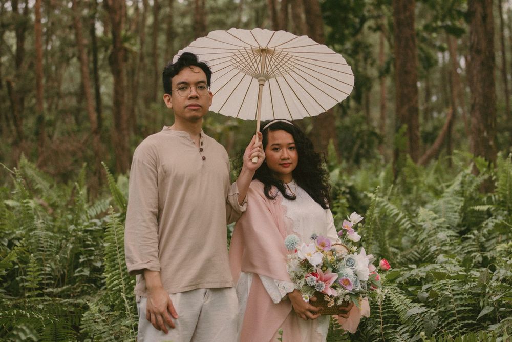 Couple poses in forest with parasol and floral bouquet, wearing soft pink and beige outfits amid lush green ferns.