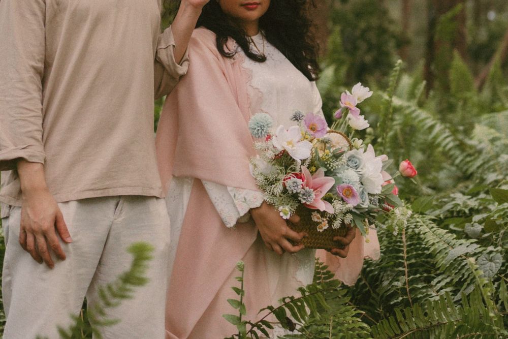 Couple in soft pink and beige attire holding a pastel floral bouquet surrounded by lush green ferns.