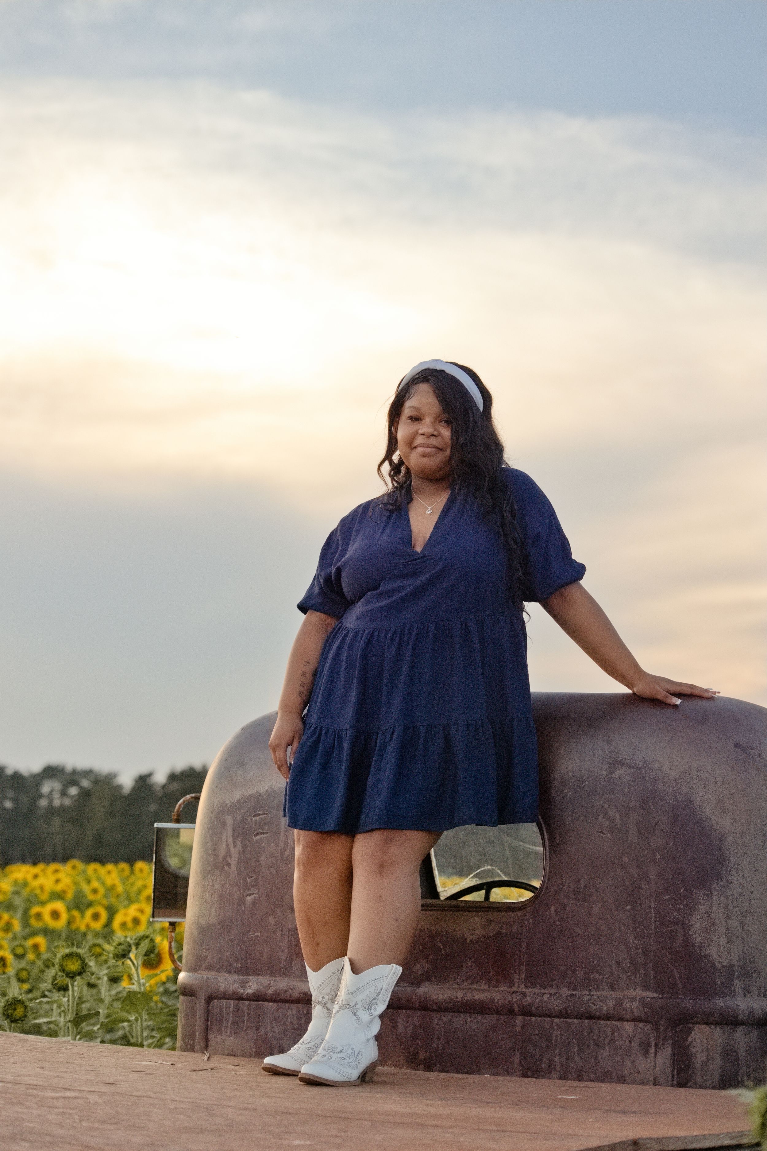 Woman in blue dress and white cowboy boots posing by rustic tractor in sunflower field at sunset.